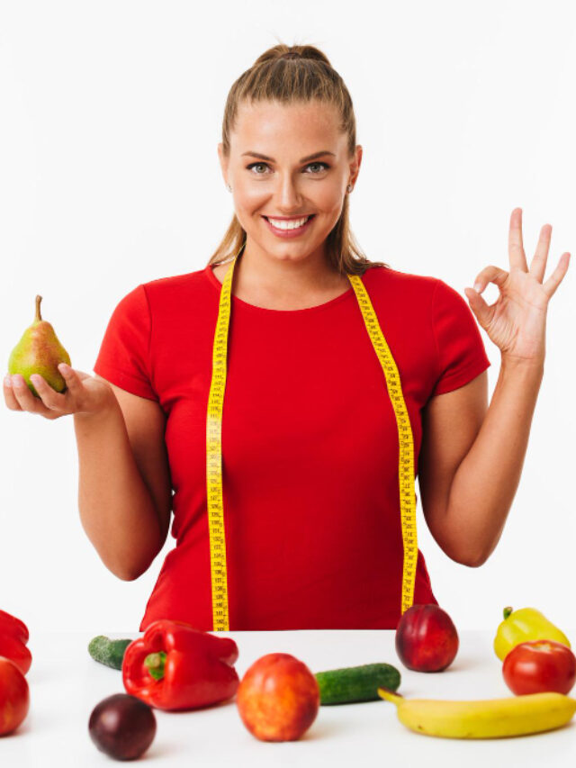 cheerful-woman-with-measuring-tape-neck-holding-pear-hand-showing-ok-gesture-while-happily-looking-camera-with-fruits-vegetables-near-white-background