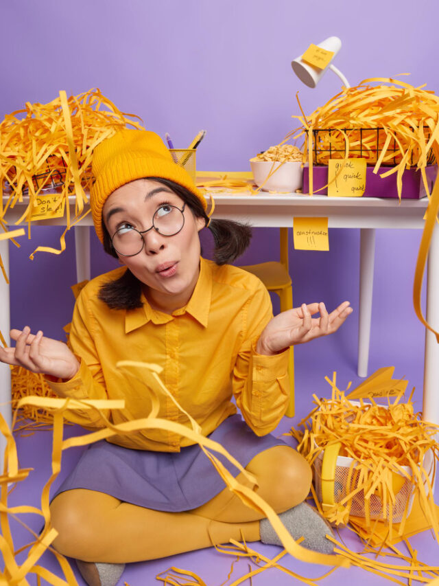 hesitant-female-model-yellow-hat-shirt-skirt-poses-crossed-legs-near-desktop-spreads-palms-has-no-clue-idea-what-first-sits-floor-own-cabinet-surrounded-py-paper-wastes